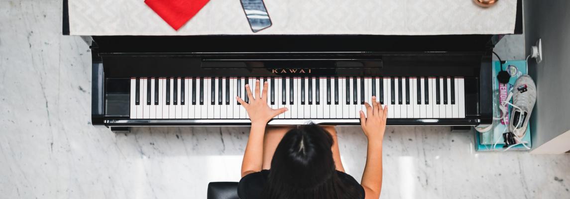 Eine dunkelhaarige Frau sitzt an einem schwarzen Klavier der Marke KAWAI, auf dem Klavier ein gerahmtes Foto eines Soldaten, ein rotes Tuch, ein Handy und drei Kerzen.